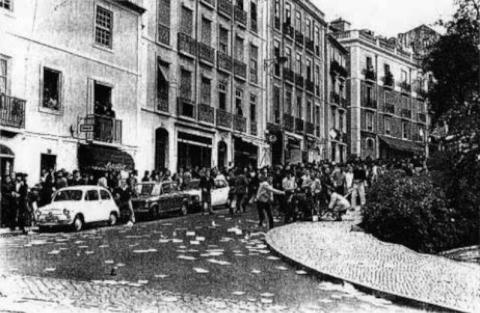 Imagem da rua de Santos-o-Velho após as primeiras cargas policiais: alguns jovens arrancam pedras da calçada para responder à violência dos pides e dos choques, vendo-se  arua juncada de comunicados.